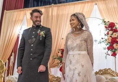 A bride and groom standing together during their wedding ceremony, with the groom in traditional dark attire and the bride in an elegant wedding dress, smiling at each other in a decorated indoor setting.