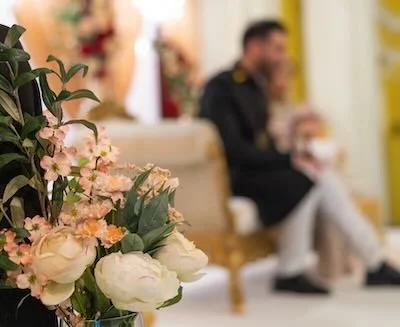A bouquet of pink, white, and peach flowers in the foreground with a blurred man and woman sitting on a sofa in the background.