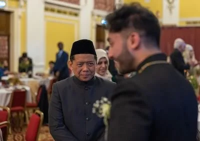 A man in a traditional black jacket and black hat looks at a man in a dark suit at a formal event in a decorated hall.