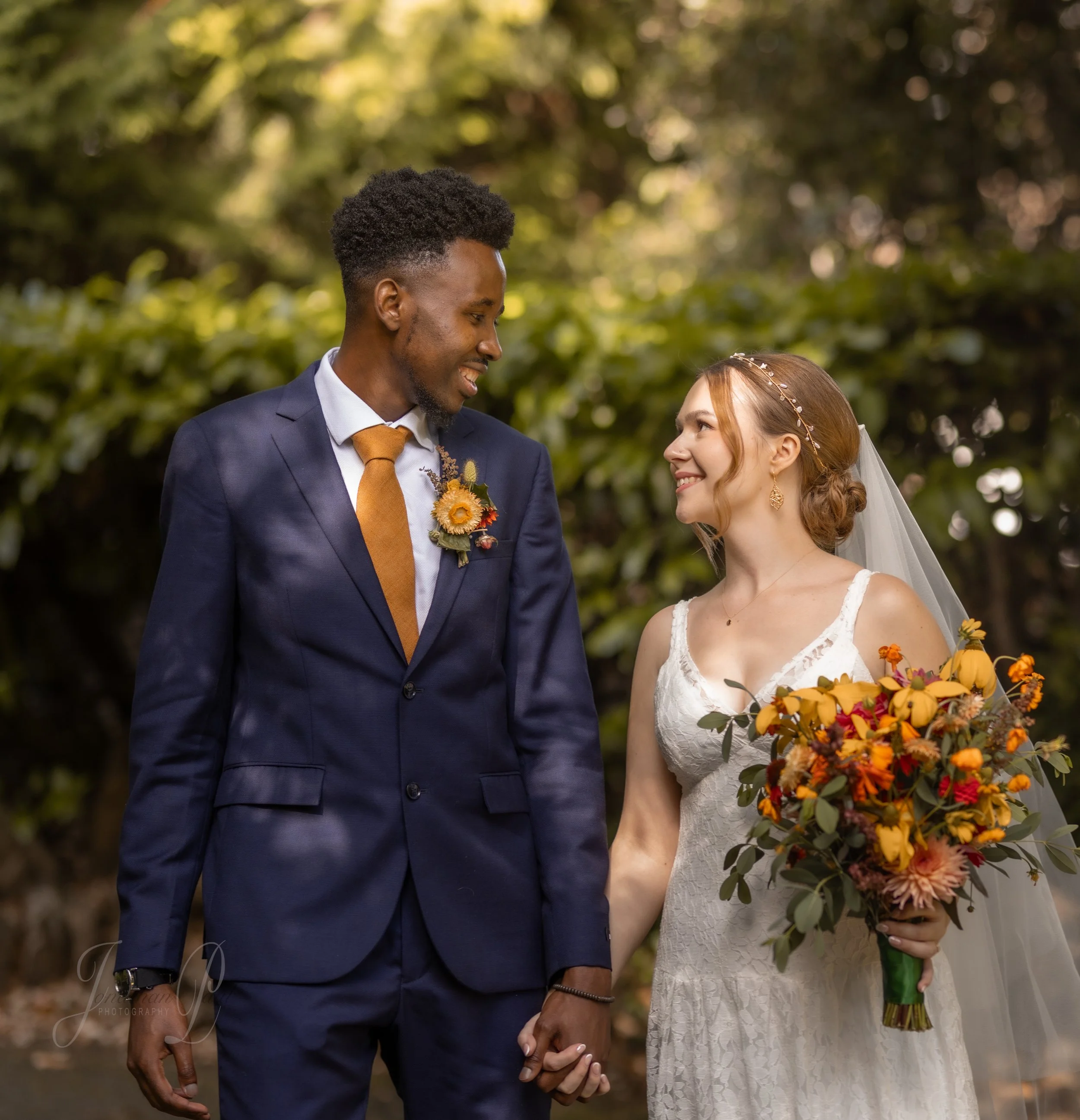 A smiling interracial couple in wedding attire holding hands outdoors in a green, sunlit park. The man wears a navy suit, white shirt, brown tie, and floral boutonniere. The woman wears a white lace dress, veil, and holds a colorful bouquet, looking