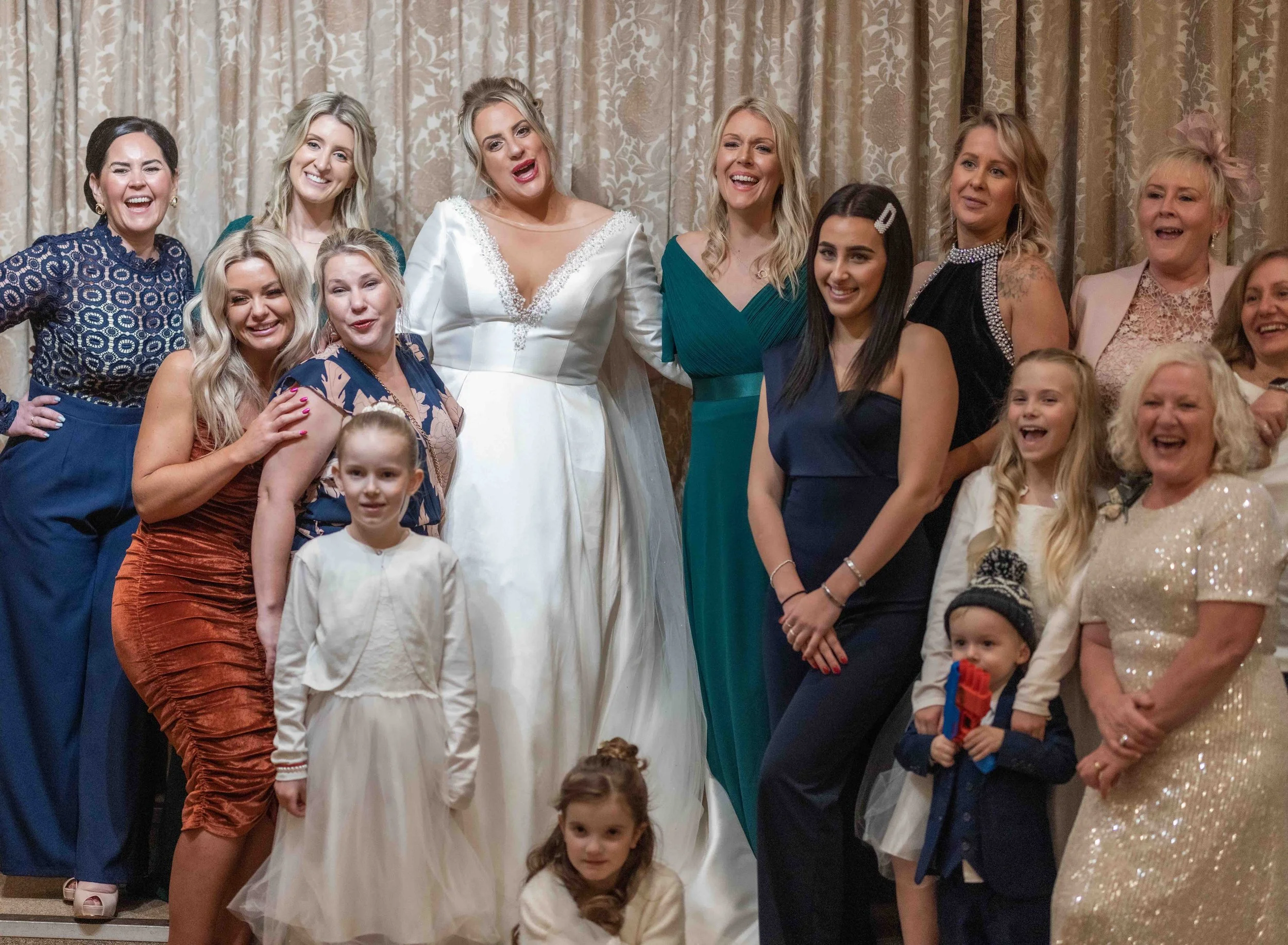 Group of women and children smiling and posing for a photo indoors, with many women wearing dresses and children in casual clothing.