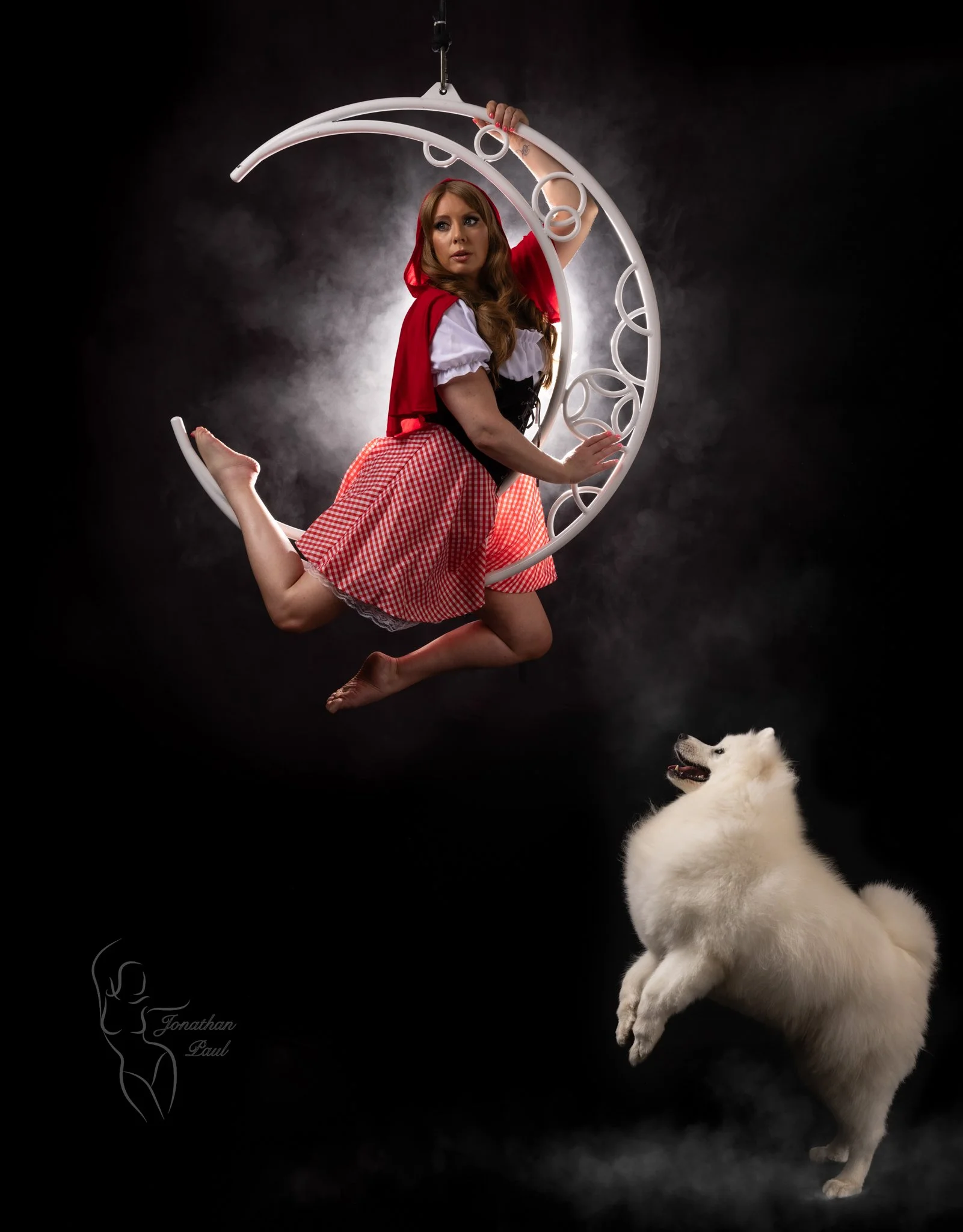 A woman dressed as Little Red Riding Hood swinging on crescent moon-shaped aerial hoop, with a white fluffy dog standing underneath against a dark background with smoke or fog.