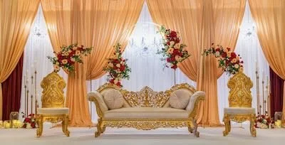 Elegant wedding stage with gold and white sofa, floral arrangements, and gold accents, set against peach curtains.