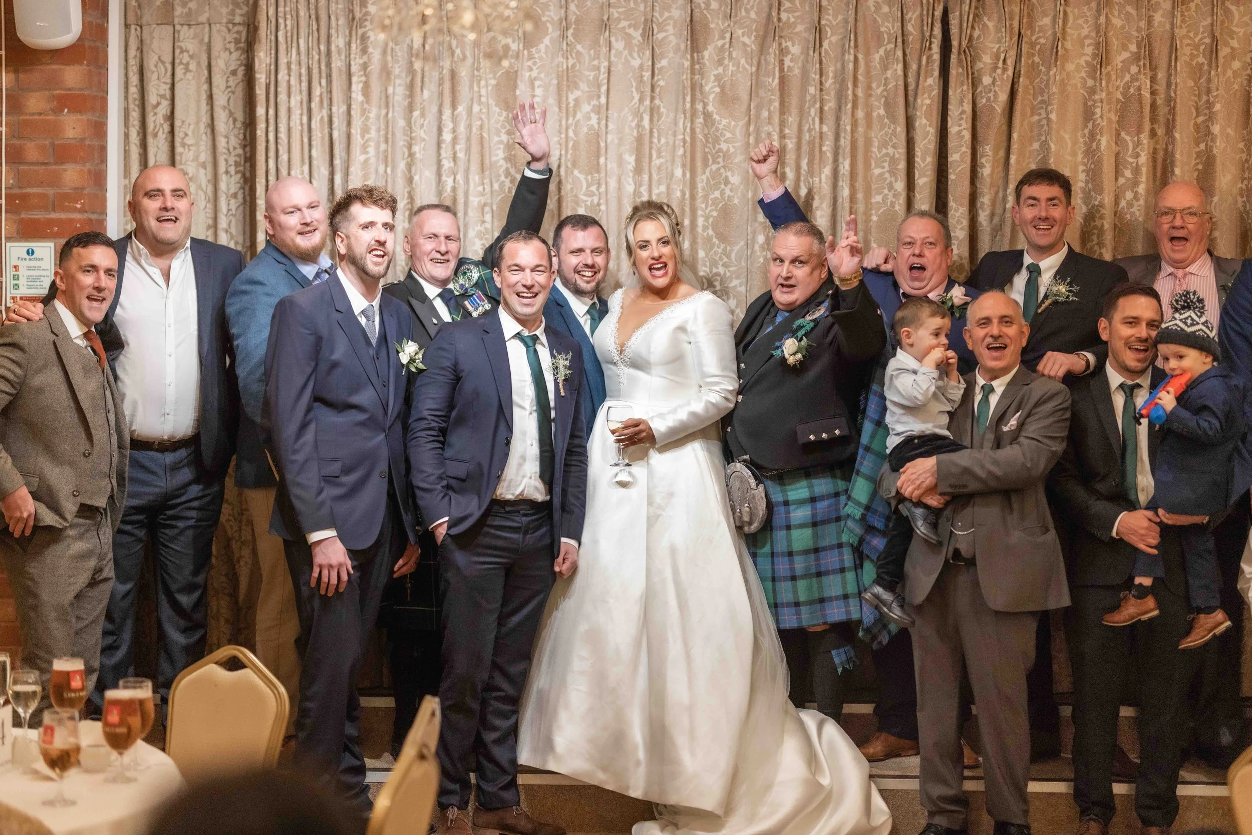 Group of wedding guests, including bride in a white gown holding a glass of wine, smiling and celebrating indoors.