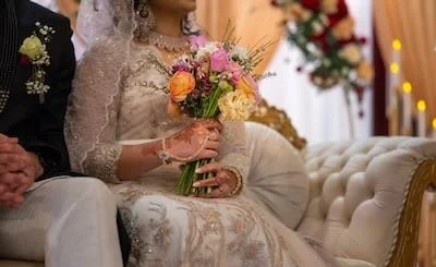 A bride in a lace wedding dress holding a bouquet of pink, yellow, and orange flowers during a wedding ceremony.