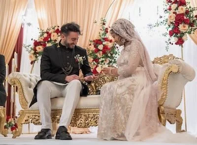 A bride and groom sitting on a decorated wedding sofa, exchanging rings, with floral arrangements in the background.