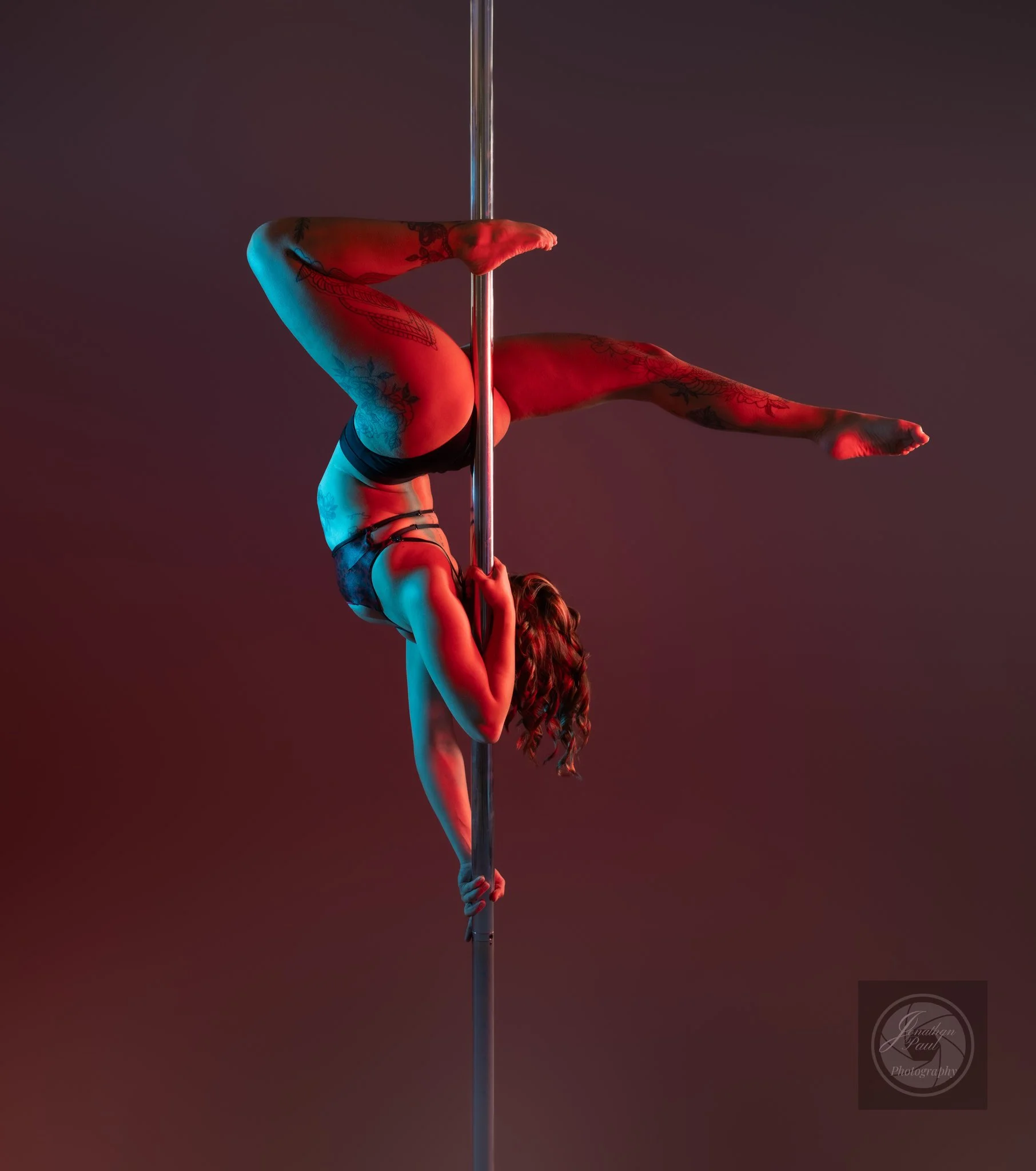 A female pole dancer performing an upside-down pose on a vertical pole, with a dark and red-lit background.