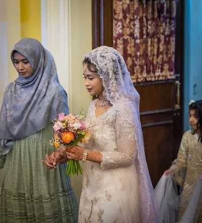 A bride holding a bouquet, wearing a lace wedding dress and veil, standing next to a woman in a hijab, with a young girl in a gold dress nearby, in a decorated indoor setting.