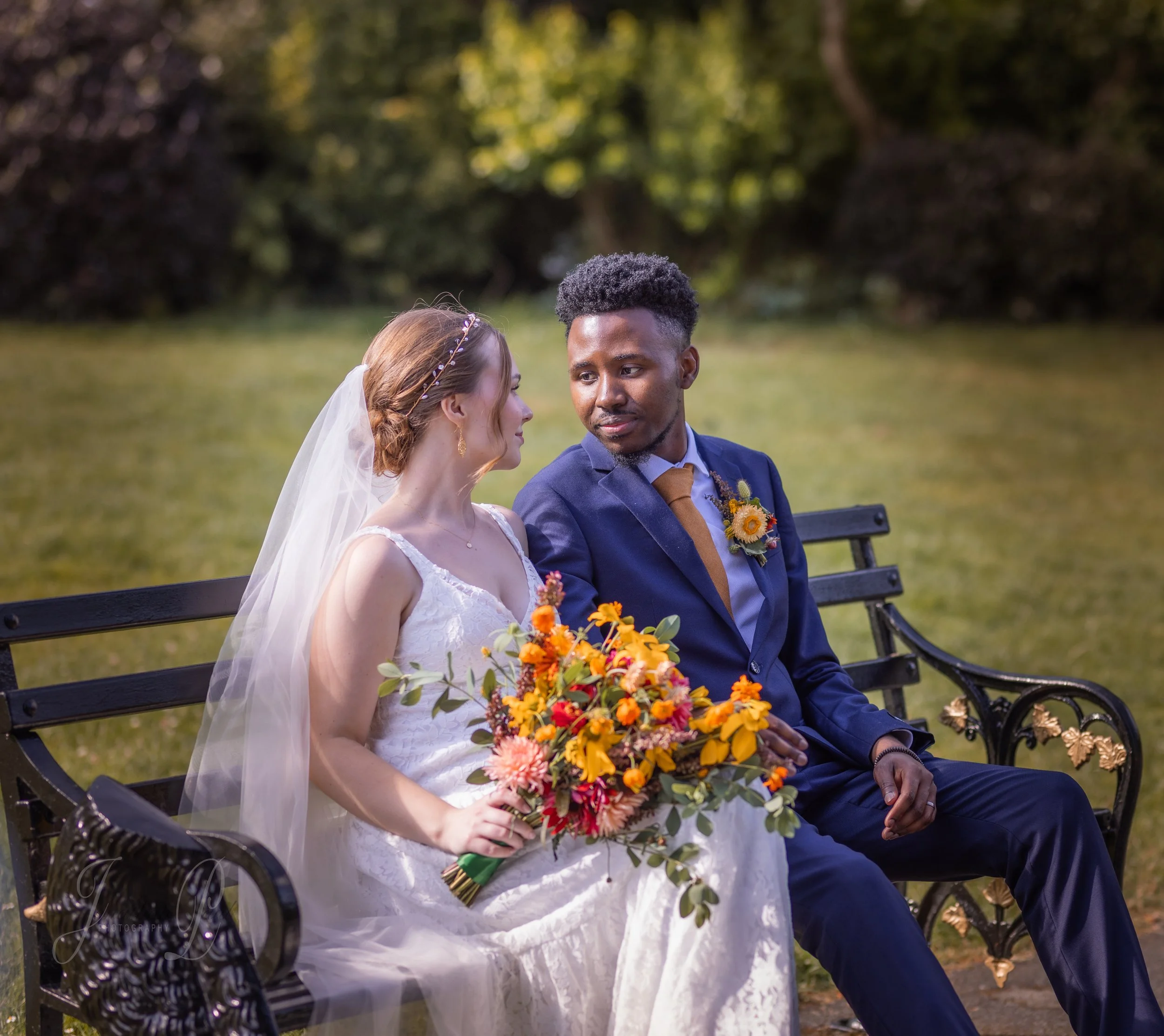 A bride and groom sitting on a park bench, gazing at each other, with the bride holding a bouquet of colorful flowers, dressed in wedding attire, outdoors with greenery in the background.