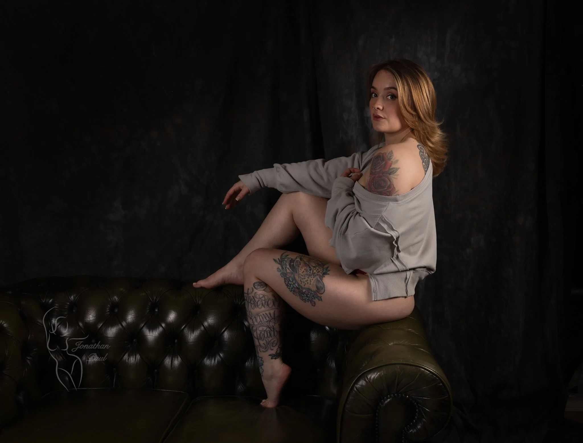 A young woman with red hair and tattoos sitting on an olive green leather couch against a dark background, wearing a beige oversized sweatshirt, one shoulder exposed, looking at the camera.