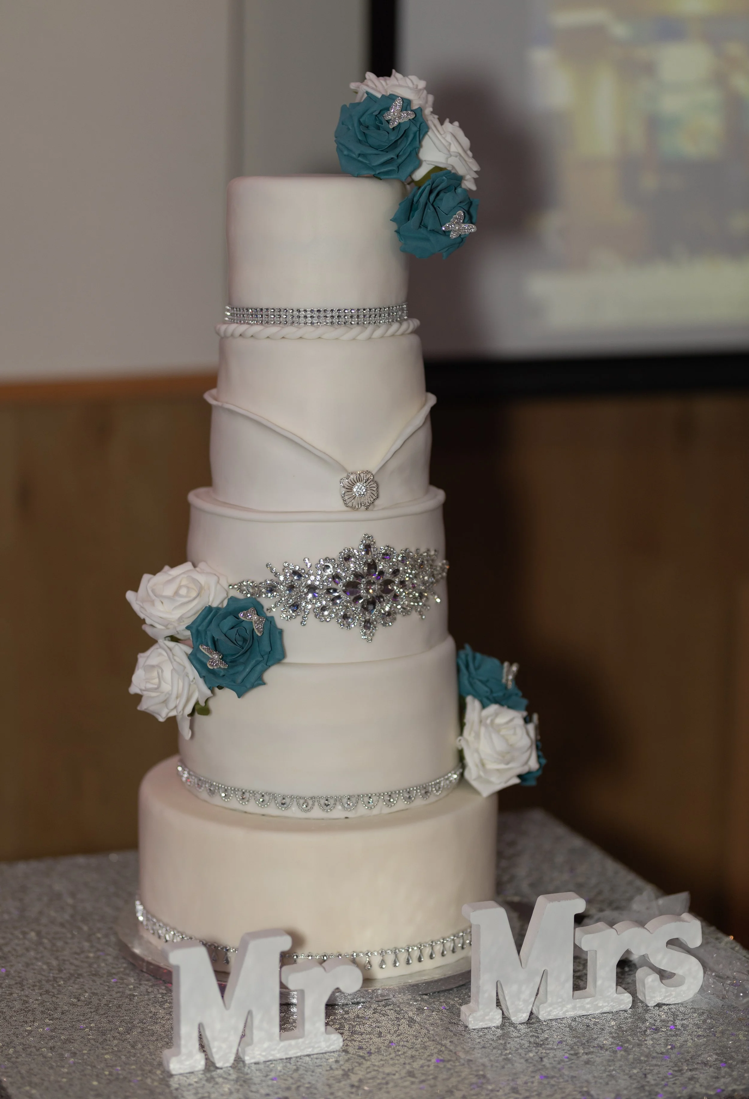 A four-tier wedding cake decorated with white and teal roses, rhinestone accents, and a "Mr" and "Mrs" sign in front of it on a glittery table.