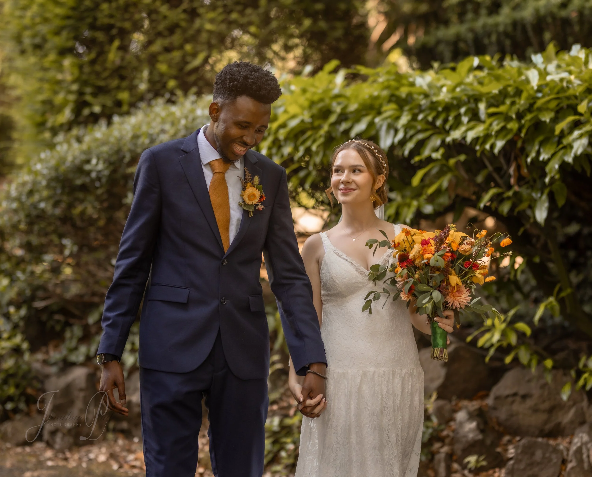A bride and groom holding hands outdoors during a wedding, with greenery and rocks in the background. The bride is holding a colorful bouquet of flowers and the groom is wearing a dark suit with a sunflower boutonniere.