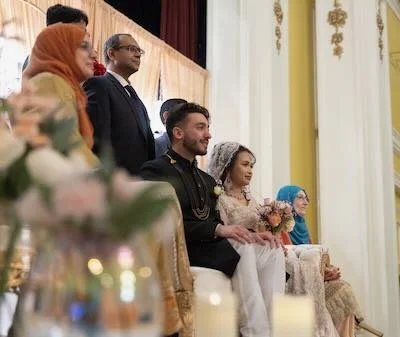 Group of people sitting on stage at a formal event or wedding, with some dressed in traditional attire.
