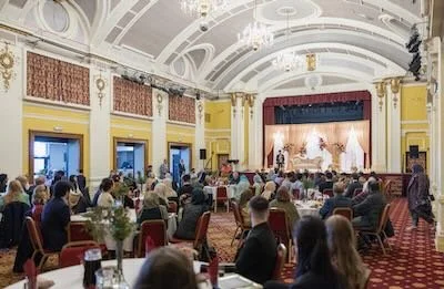 People attending a formal event or conference in a large, ornate ballroom with a stage. Attendees are seated at round tables with white tablecloths, and the room features yellow walls, decorative moldings, and a high, arched ceiling.