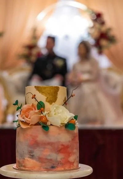 A two-layer wedding cake decorated with flowers in front of a blurred bride and groom during their wedding reception.