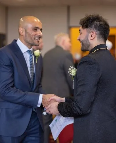 Two men in formal attire shaking hands at a formal event or wedding, smiling, with other guests in the background.