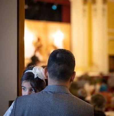A young girl looks over a man's shoulder in an indoor setting, possibly a church or event hall, with warm lighting and blurred background.