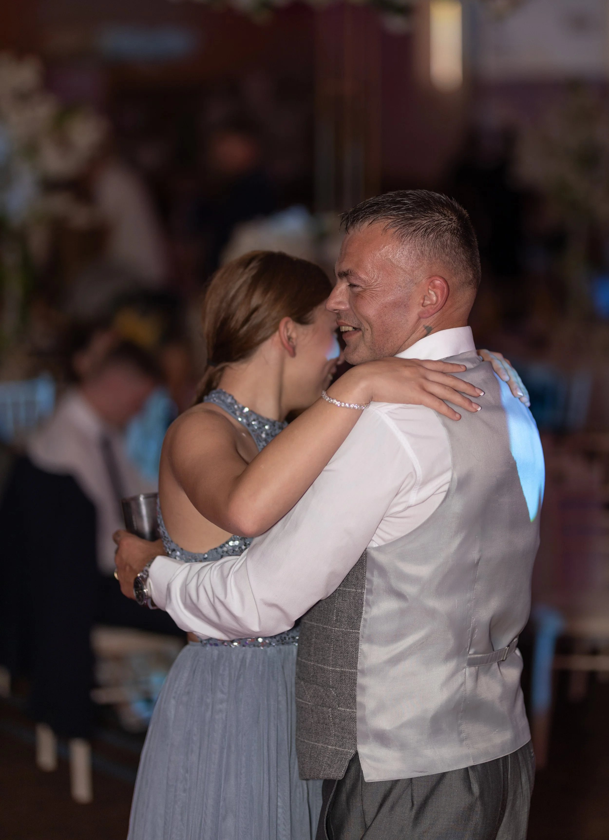 A couple dancing closely and smiling at a wedding reception, with blurred guests in the background.