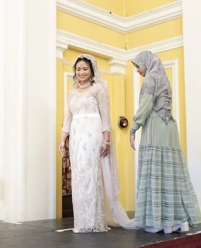 A woman in a white lace wedding dress with a veil stands smiling, while another woman in a plaid skirt and gray hijab adjusts her dress in a yellow room with white molding.