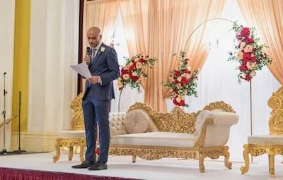 A man in a suit standing on a stage, reading from papers, with a decorated backdrop and a fancy sofa.