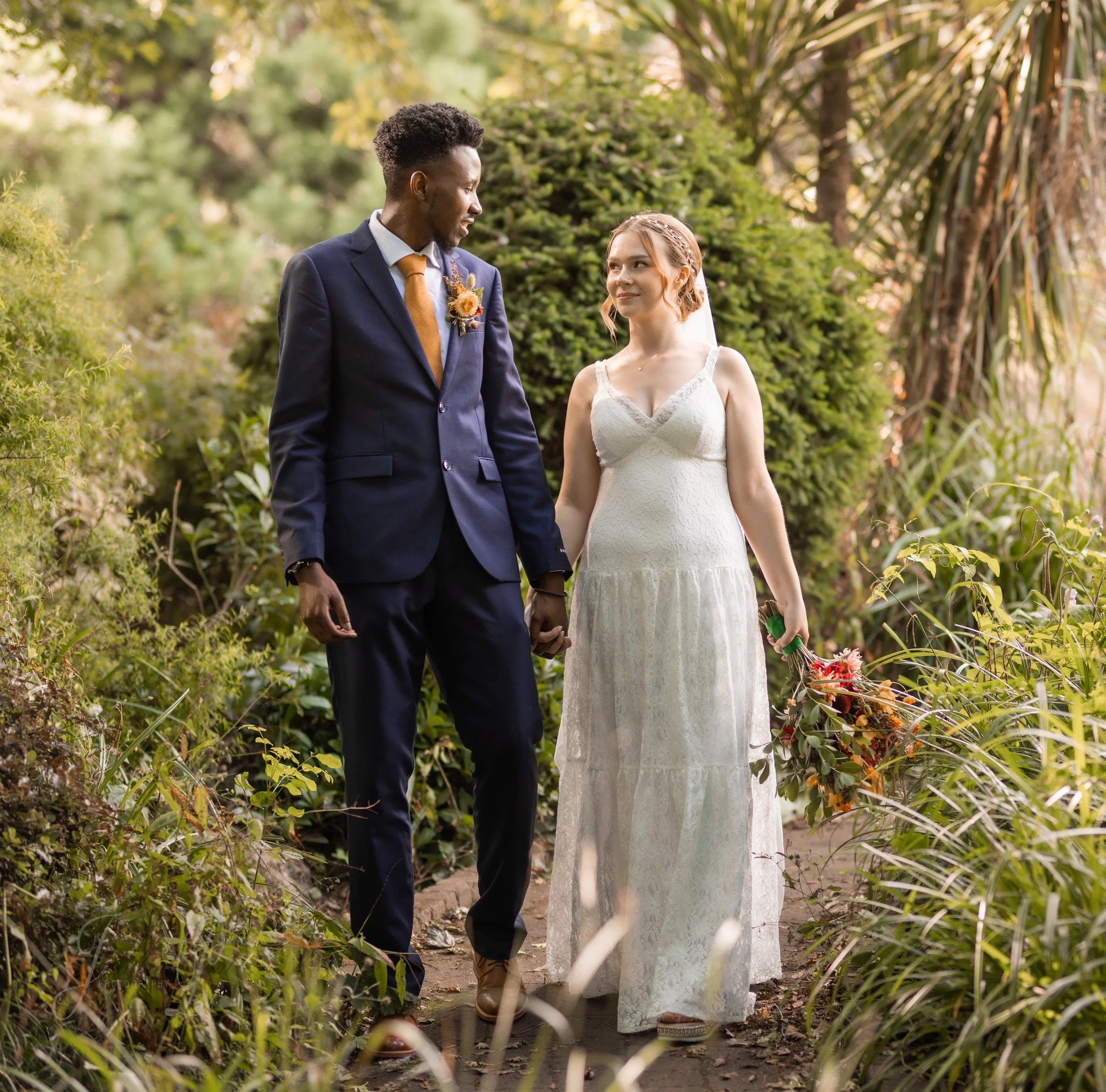 A couple holding hands, walking outdoors in a lush garden, dressed in wedding attire, with the man in a navy suit and the woman in a white lace dress, holding a bouquet.
