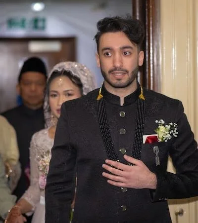 Man in traditional black attire with a boutonniere, standing in front of a woman in bridal attire, at a wedding ceremony.