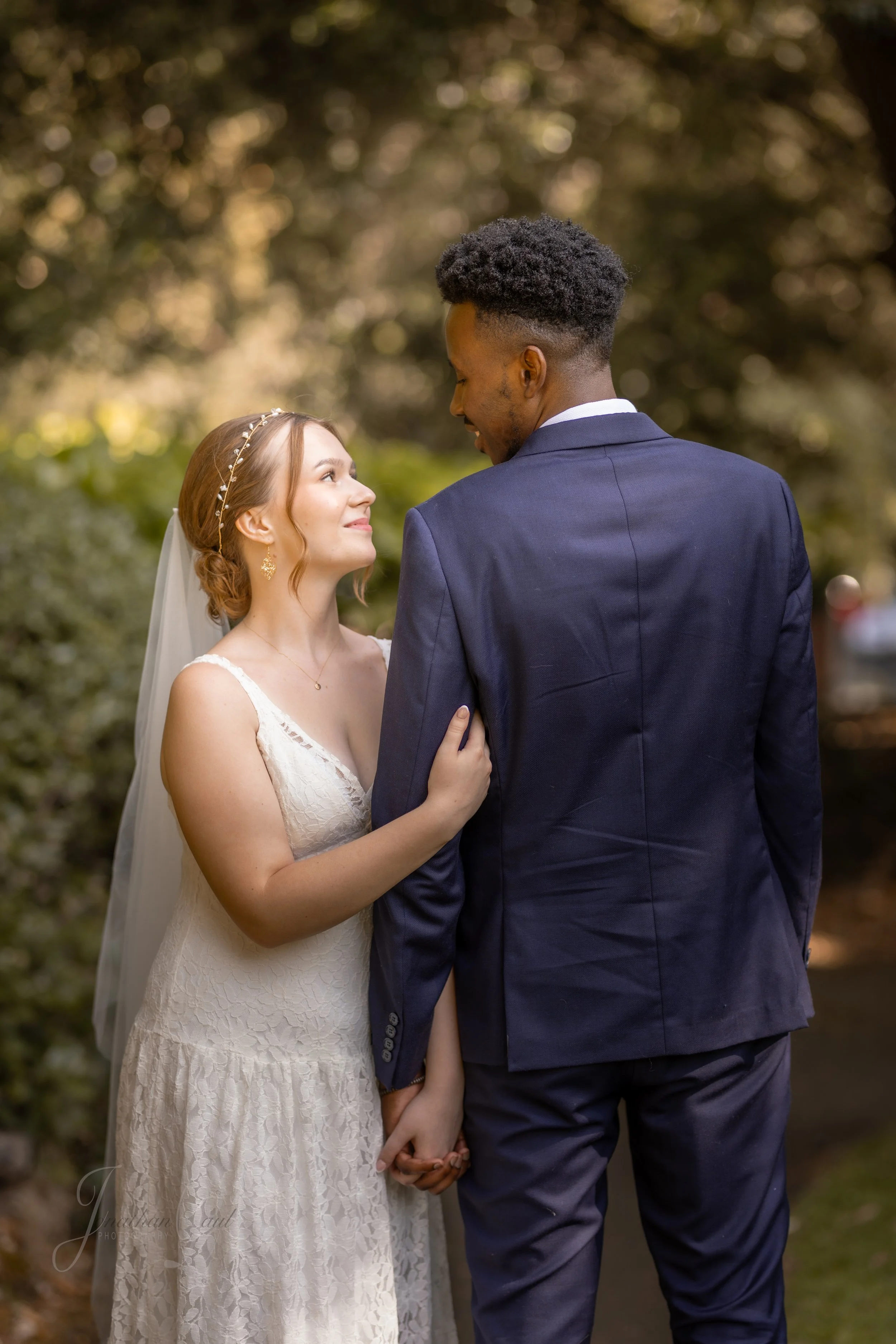A bride and groom standing close, smiling, and looking into each other's eyes outdoors with blurred trees in the background.
