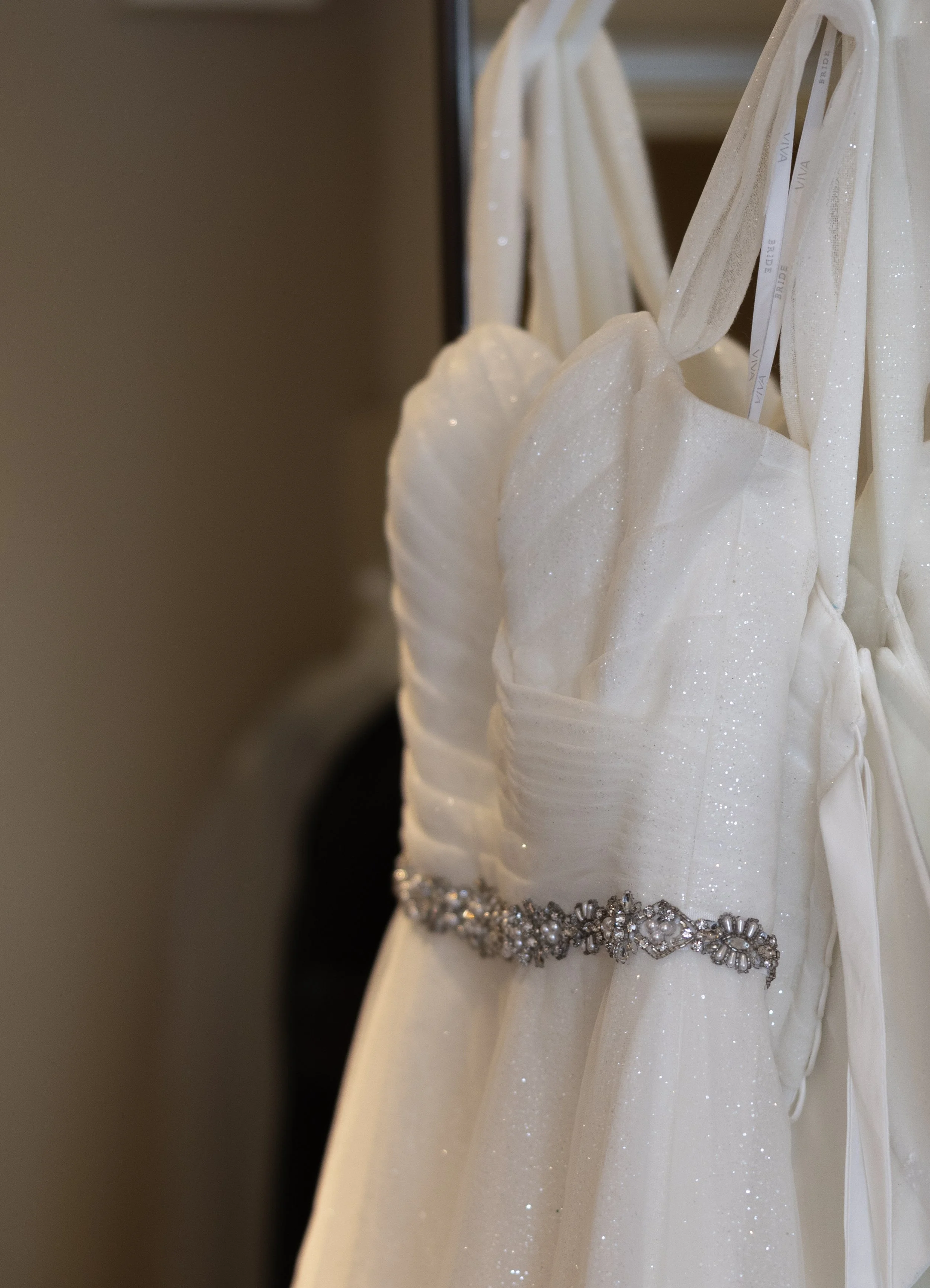White wedding dress with a sparkling fabric and a decorative rhinestone belt hanging on a rack.