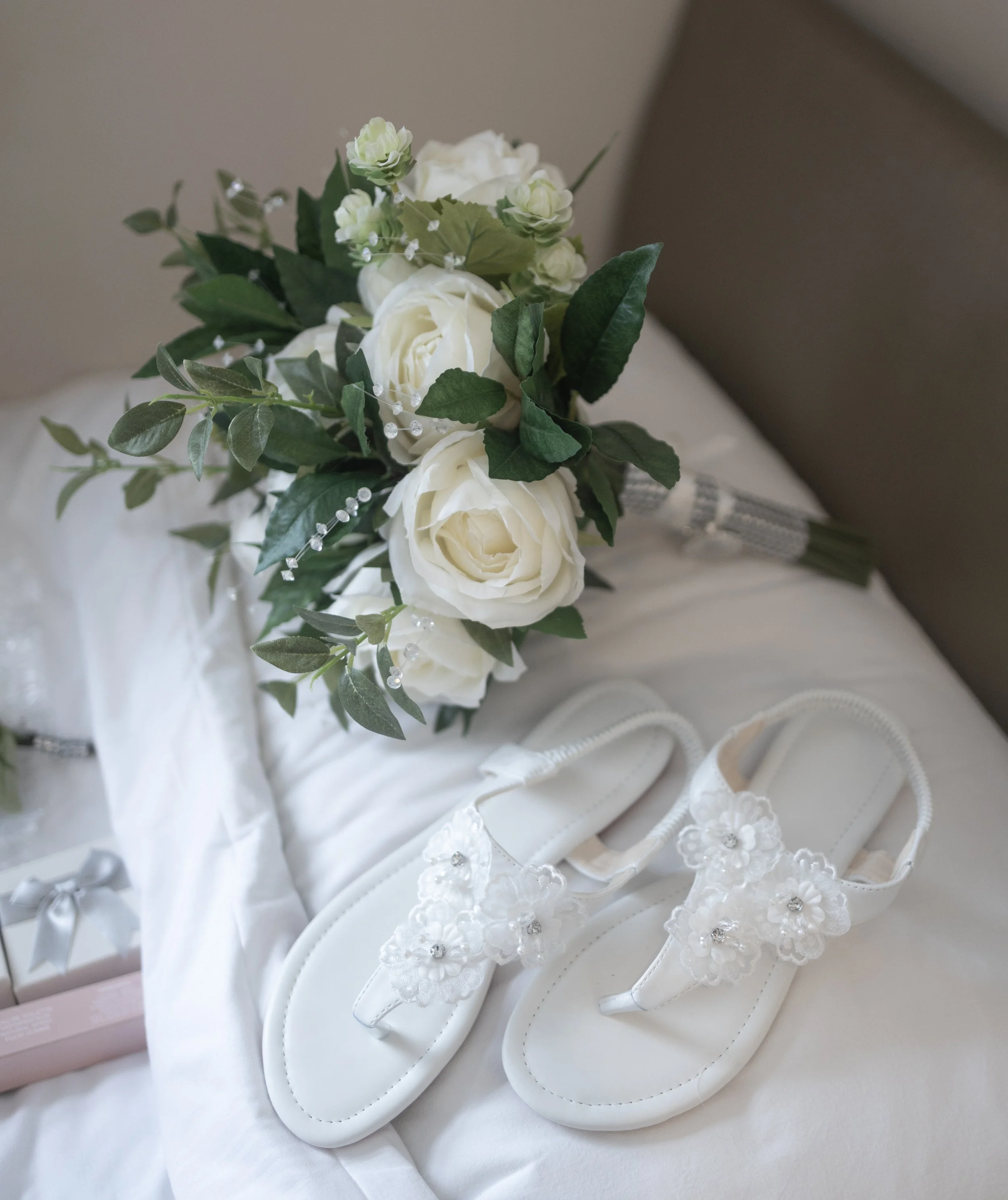 White wedding bouquet with roses and greenery, white sandals with floral embellishments, and a small gift box on a bed.