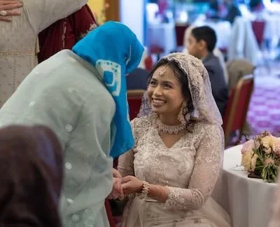 A woman in traditional wedding attire smiling and holding hands with another woman wearing a blue hijab at a wedding reception.