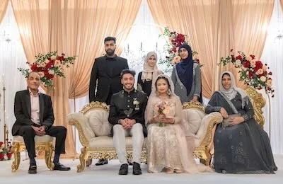 A group of seven people posing on a decorated stage for a celebration, possibly a wedding. The group includes a bride and groom sitting in the center, surrounded by family members, with floral arrangements and elegant drapery in the background.