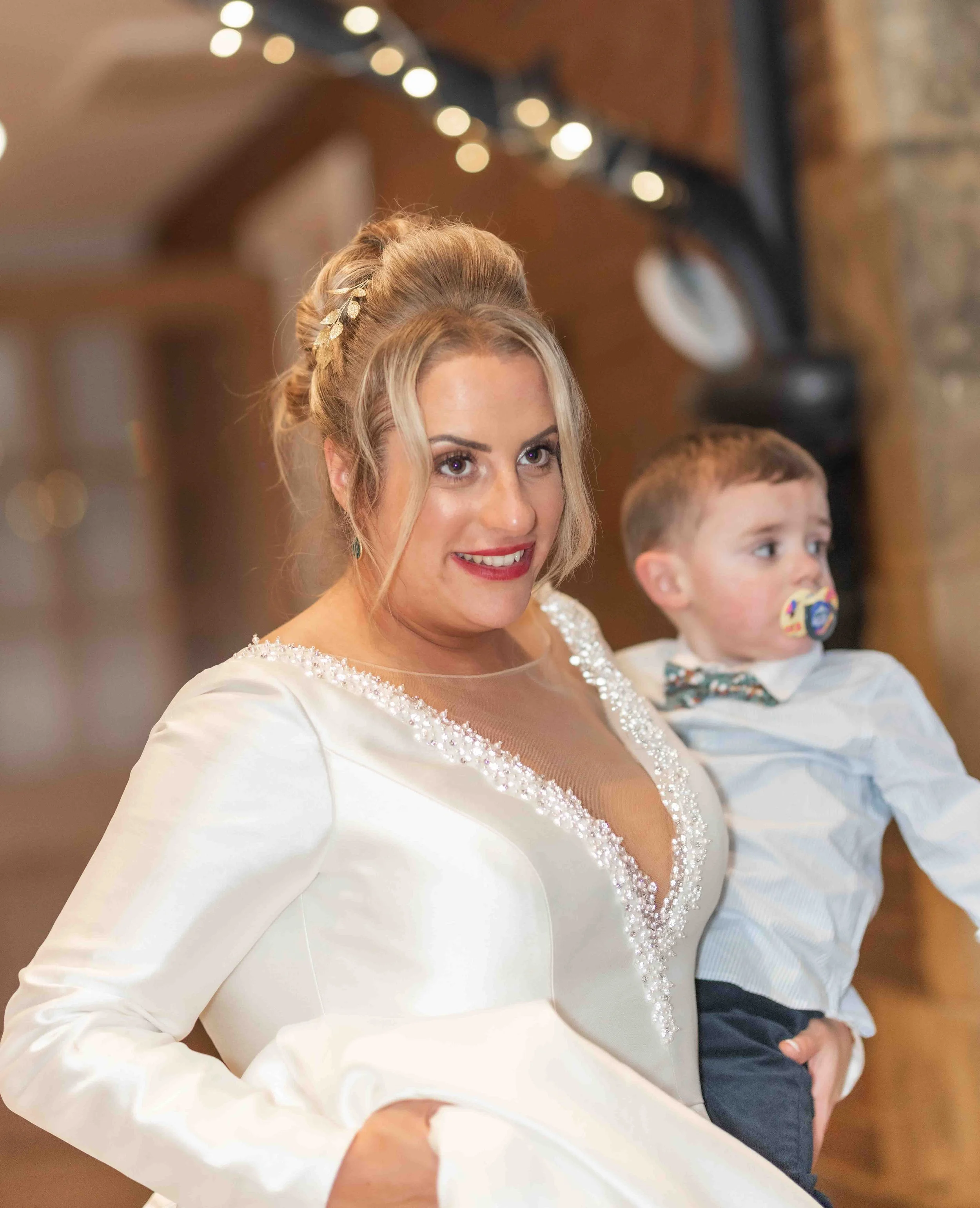 A woman in a white dress holding a young boy, both smiling, in a warmly lit indoor setting.