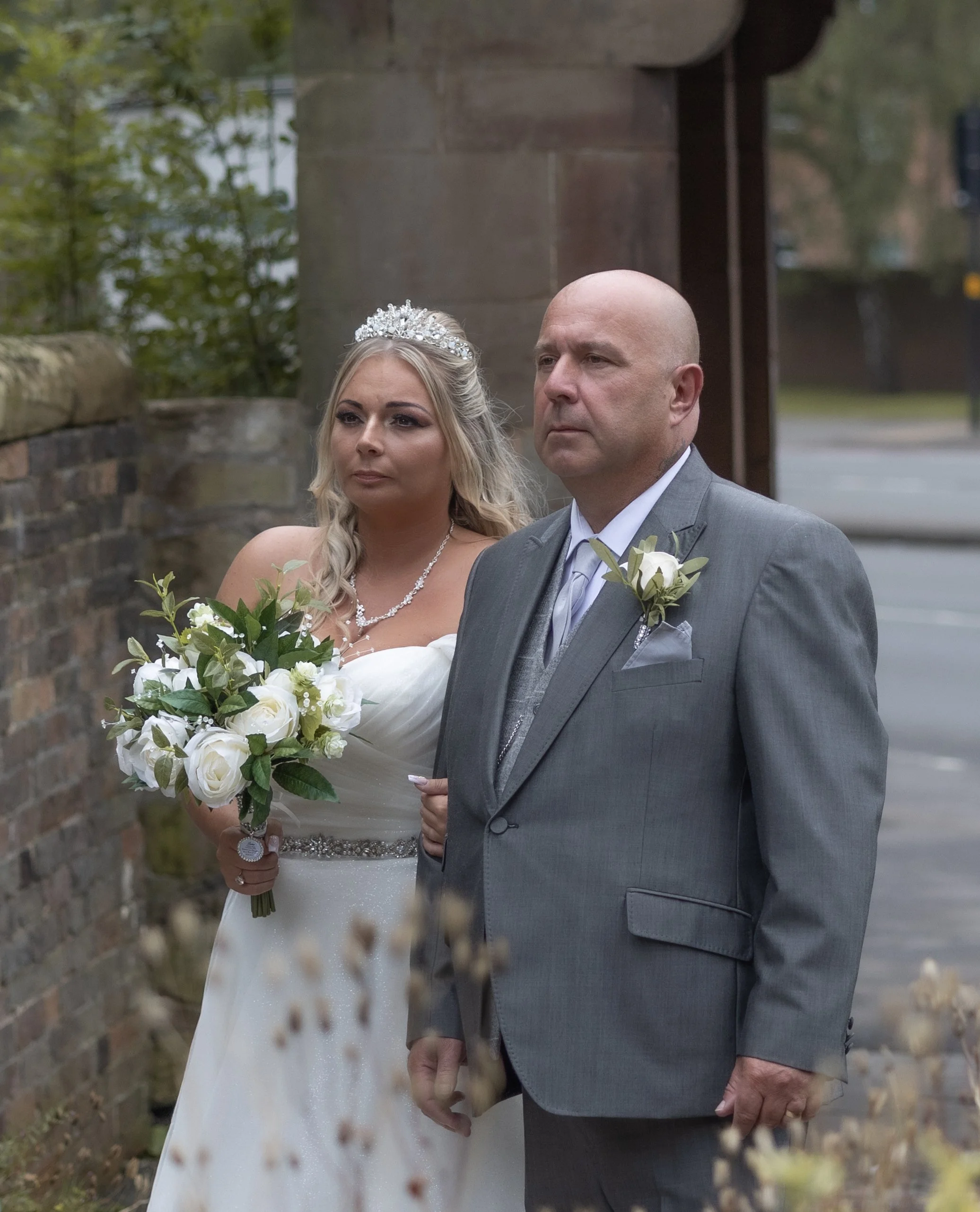 A bride in a white wedding dress holding a bouquet, walking arm in arm with a man in a gray suit, during a wedding ceremony outdoors near a brick wall.
