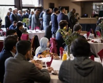 People attending a formal event or conference in a banquet hall with round tables, food, and drinks.