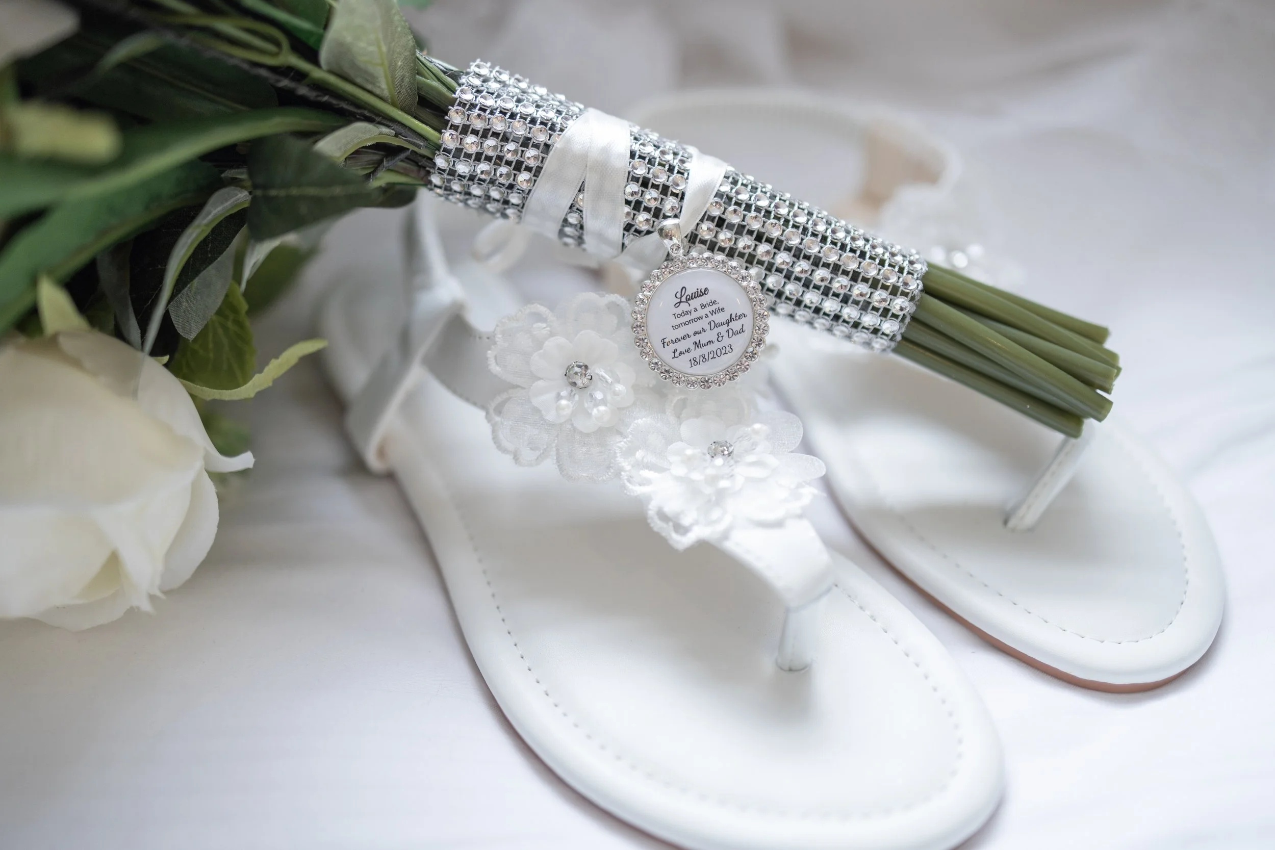 White wedding sandals with white floral accents, a bouquet with green leaves, and a rhinestone bracelet with a personalized pendant resting on the bouquet.