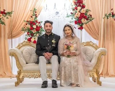 A newlywed couple sits on an ornate cream-colored sofa with gold accents, surrounded by pink and red floral arrangements and peach curtains at a wedding ceremony.