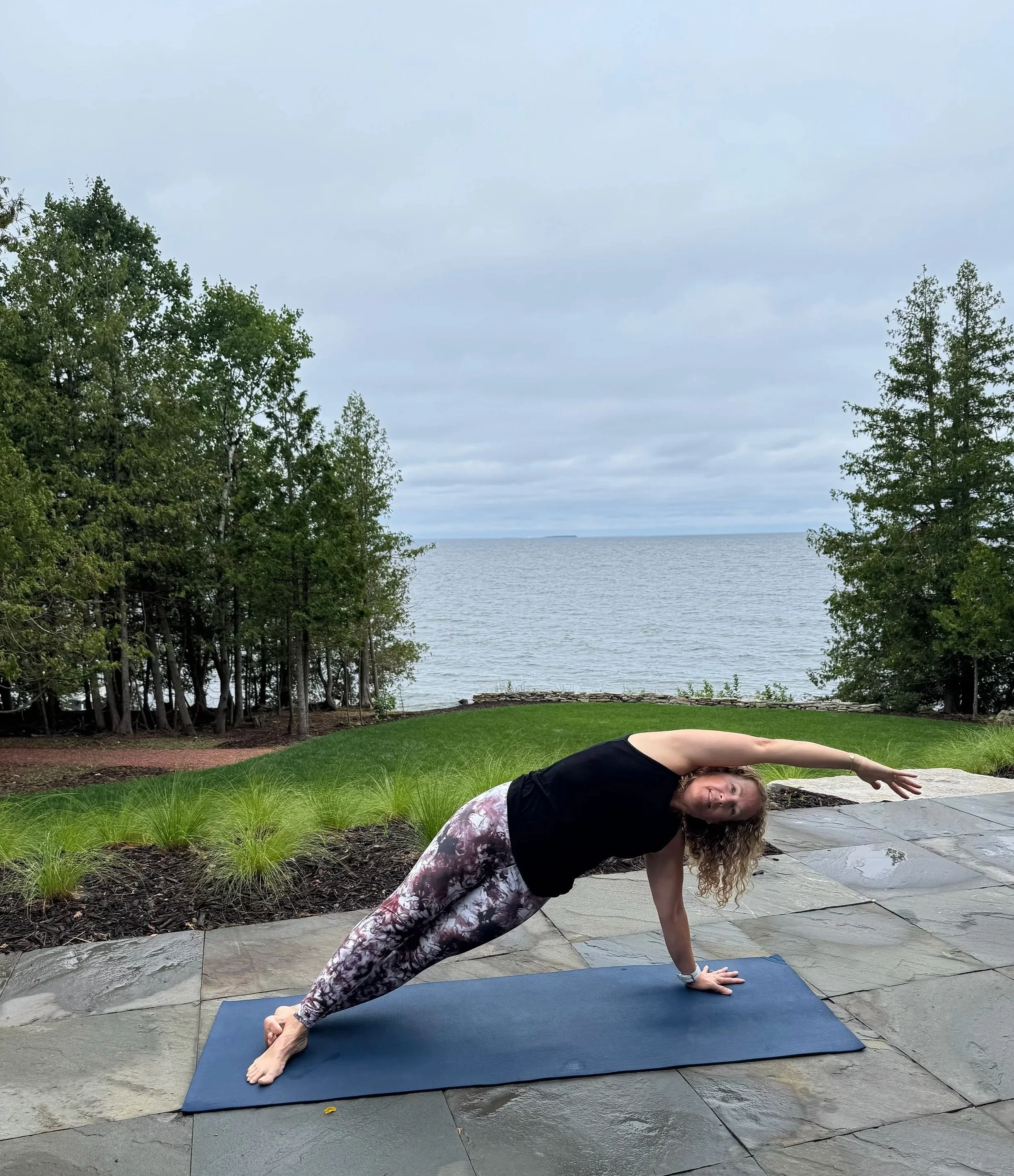 A woman practicing yoga outdoors on a mat near a body of water, with greenery and trees surrounding her; she is in a side plank pose, wearing patterned leggings and a black top.