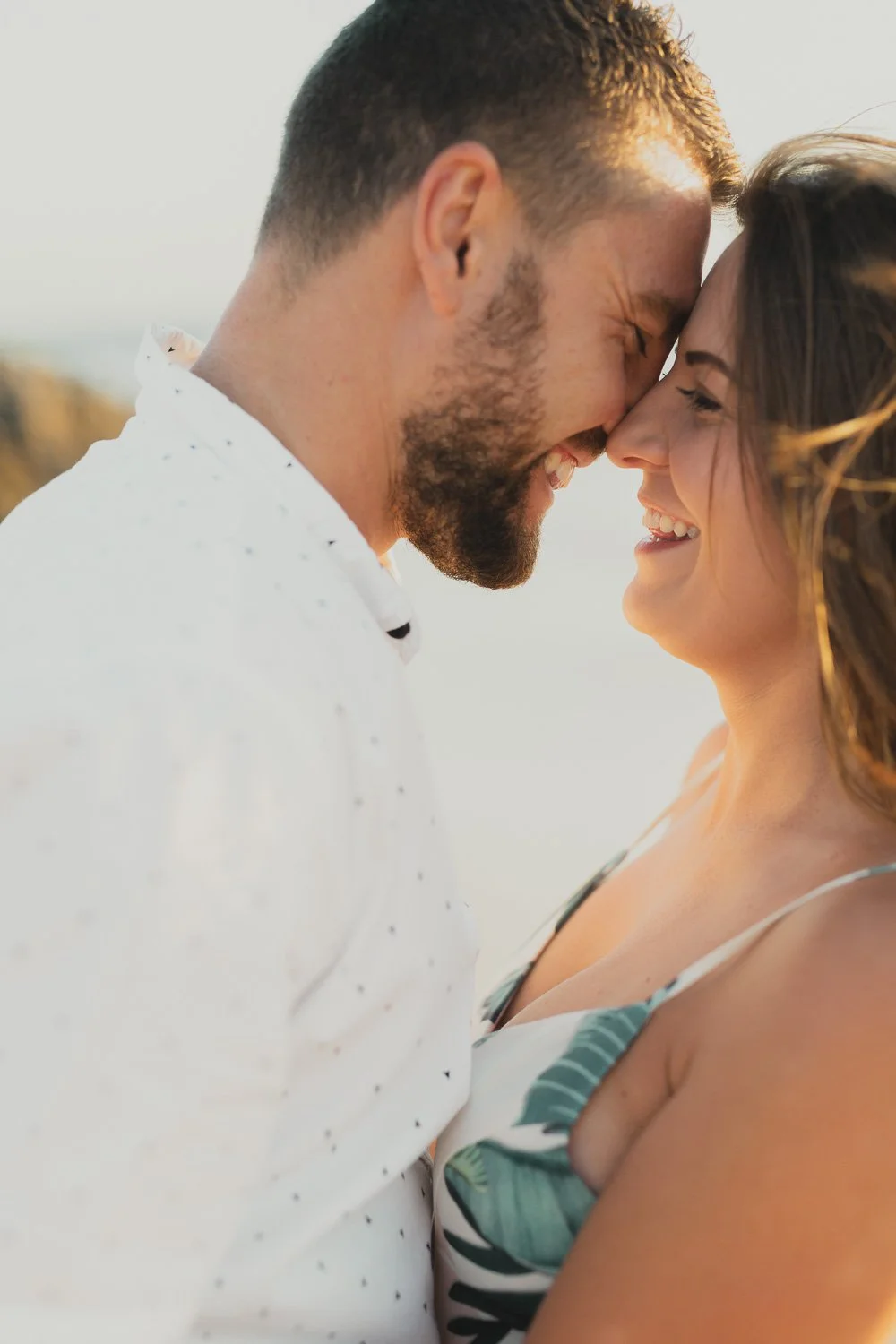 A smiling man and woman with their foreheads touching, outdoors during sunset, close-up of their faces with warm light.