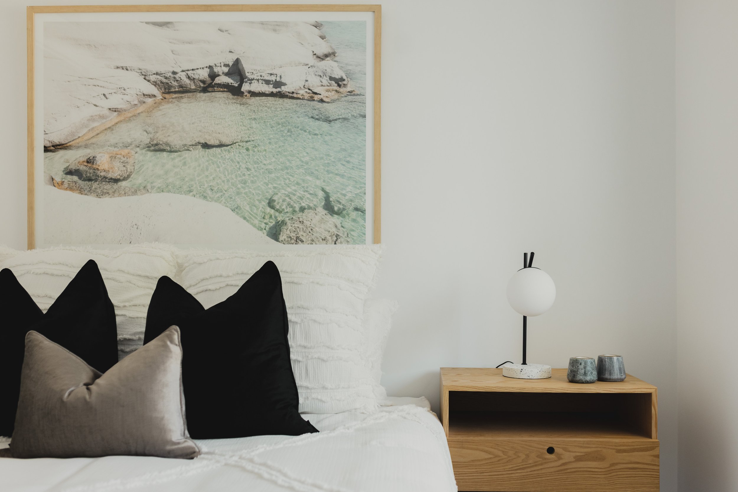 A bedroom corner with a bed, black and gray pillows, a wooden bedside table, a modern lamp, and a framed aerial photograph of a coastal landscape.