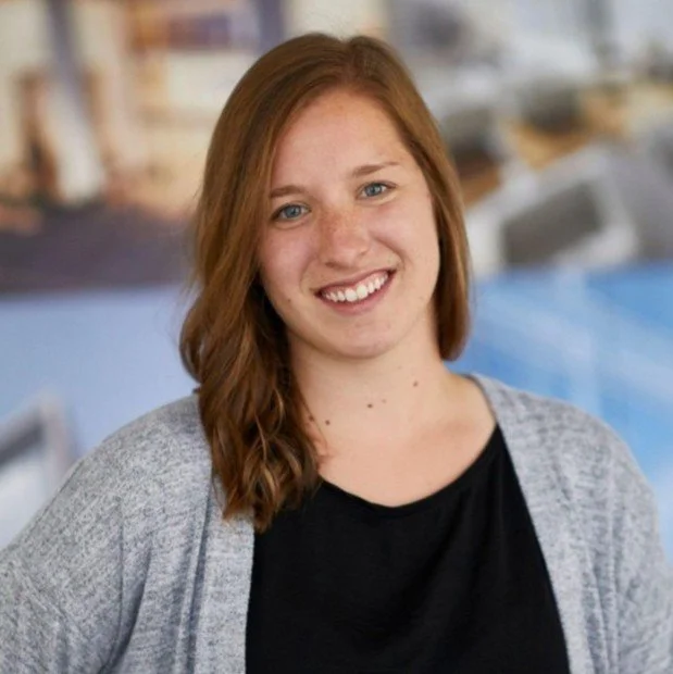 professional headshot of a smiling woman with short brown hair, black tank top, and gray cardigan, on a blue background