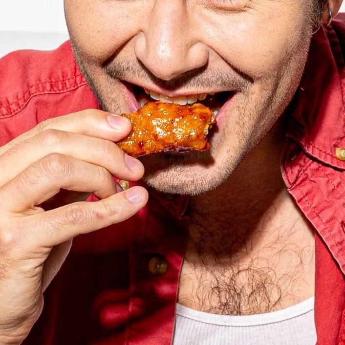 Close-up of a man in a red shirt eating a glazed chicken wing.