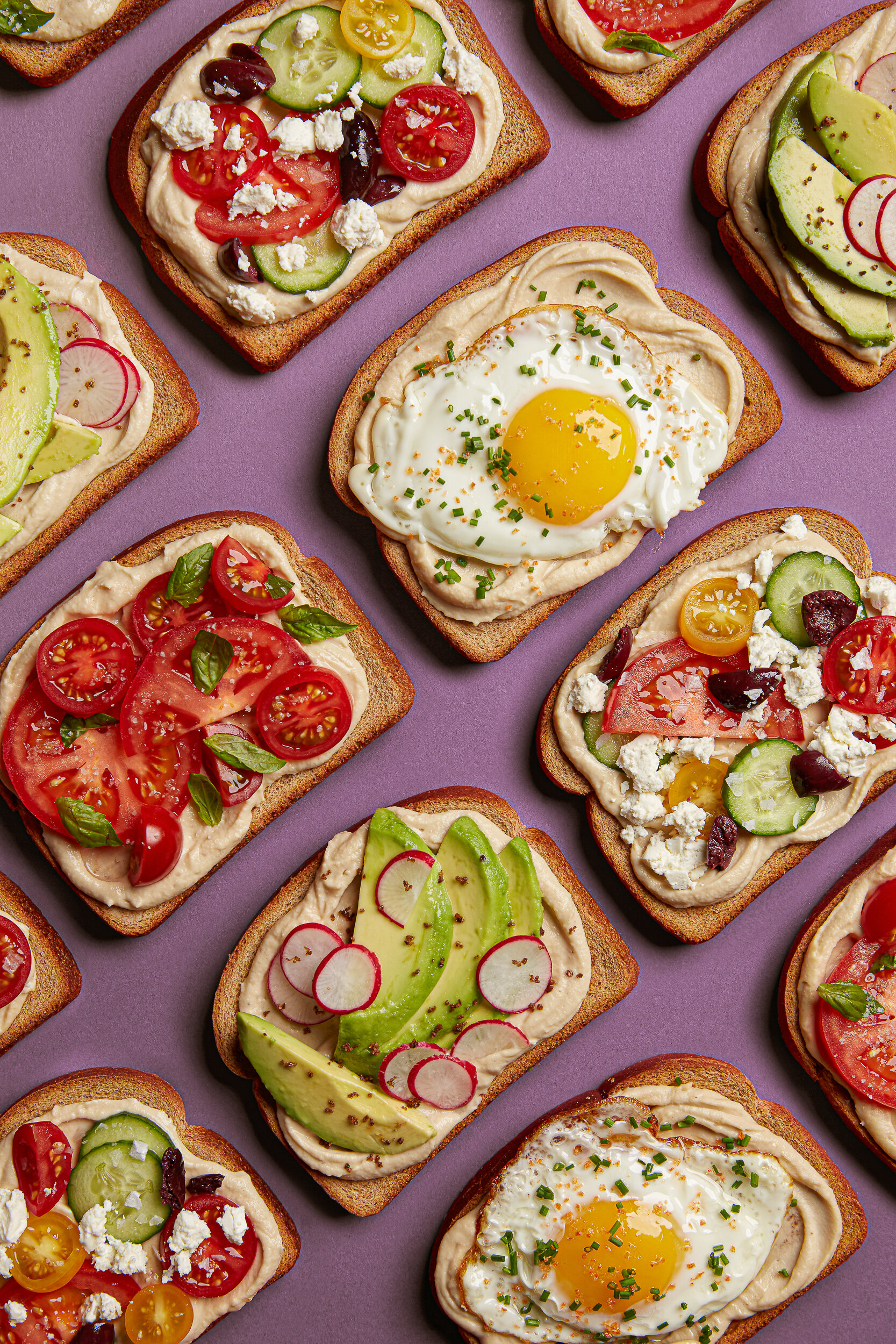 Variety of open-faced sandwiches on a purple background, featuring toppings like avocado, sliced radishes, tomatoes, fried eggs, hummus, olives, and feta cheese.