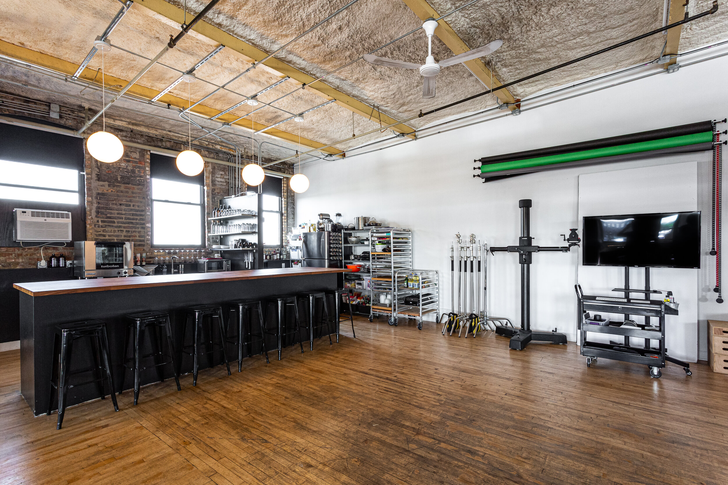 Modern photography studio interior with industrial design, featuring a long counter with bar stools, overhead lighting, camera equipment, a television, and backdrop rollers on wall.