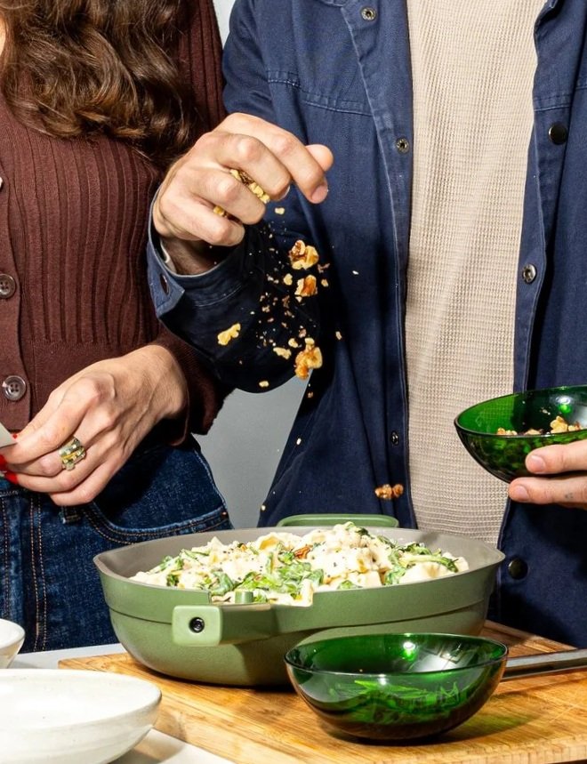 Huge Galdones photograph of people gathering around a table with a green bowl of pasta and green salad bowls, with one person sprinkling crumbled toppings onto the dish.