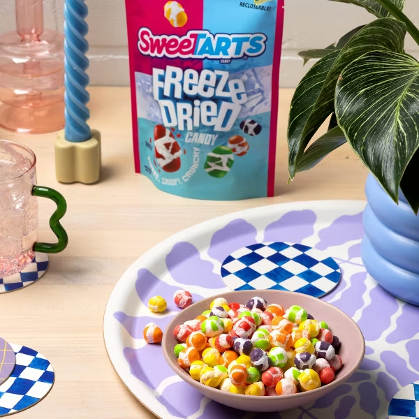Huge Galdones photograph for Ferrara Candy Sweetarts A bowl of colorful candy beads on a decorated plate with a box of SweetTarts Freeze Dried Candy in the background, a green straw in a pink drink, and a potted plant on a wooden surface.