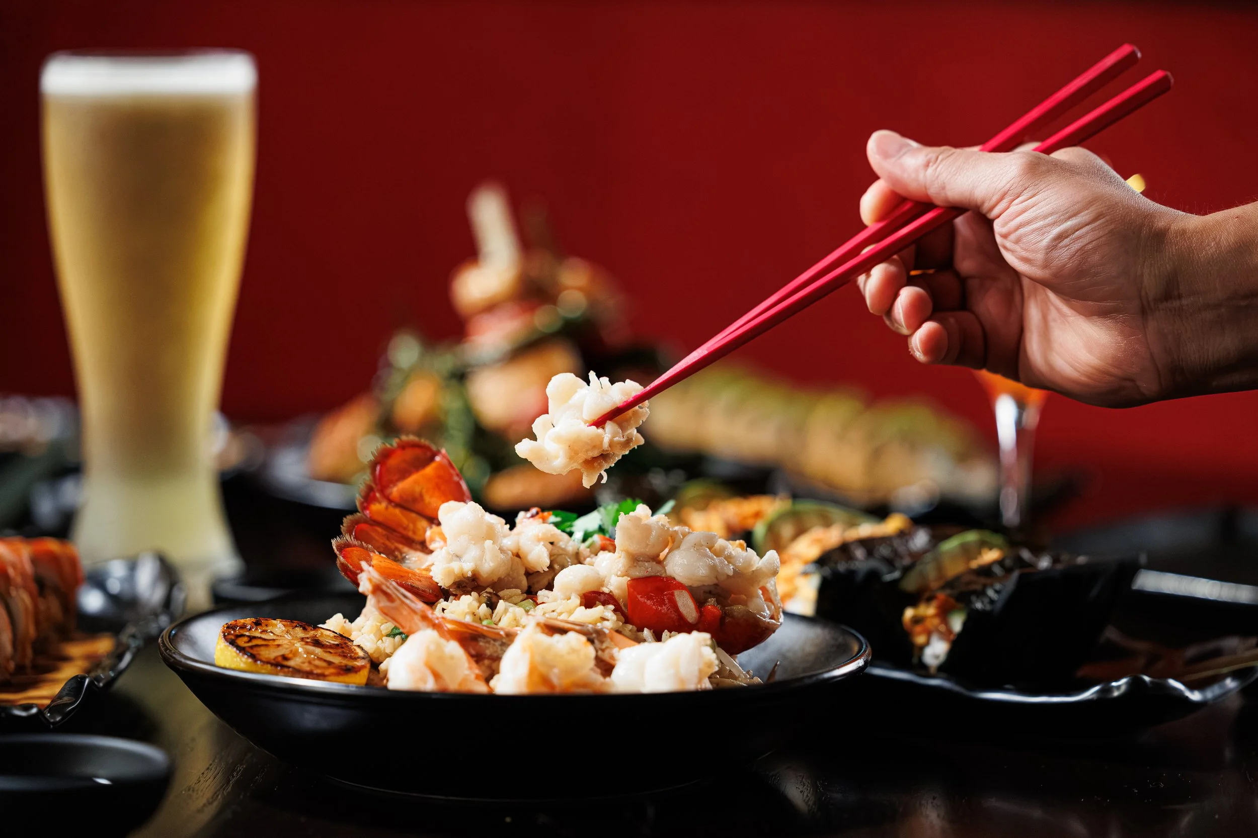 Huge Galdones food and lifestyle photography for P.F. Chang's showing a close-up of a hand holding red chopsticks picking up sushi on a black plate, with a tall glass of beer in the background, surrounded by various sushi pieces on a black platter.