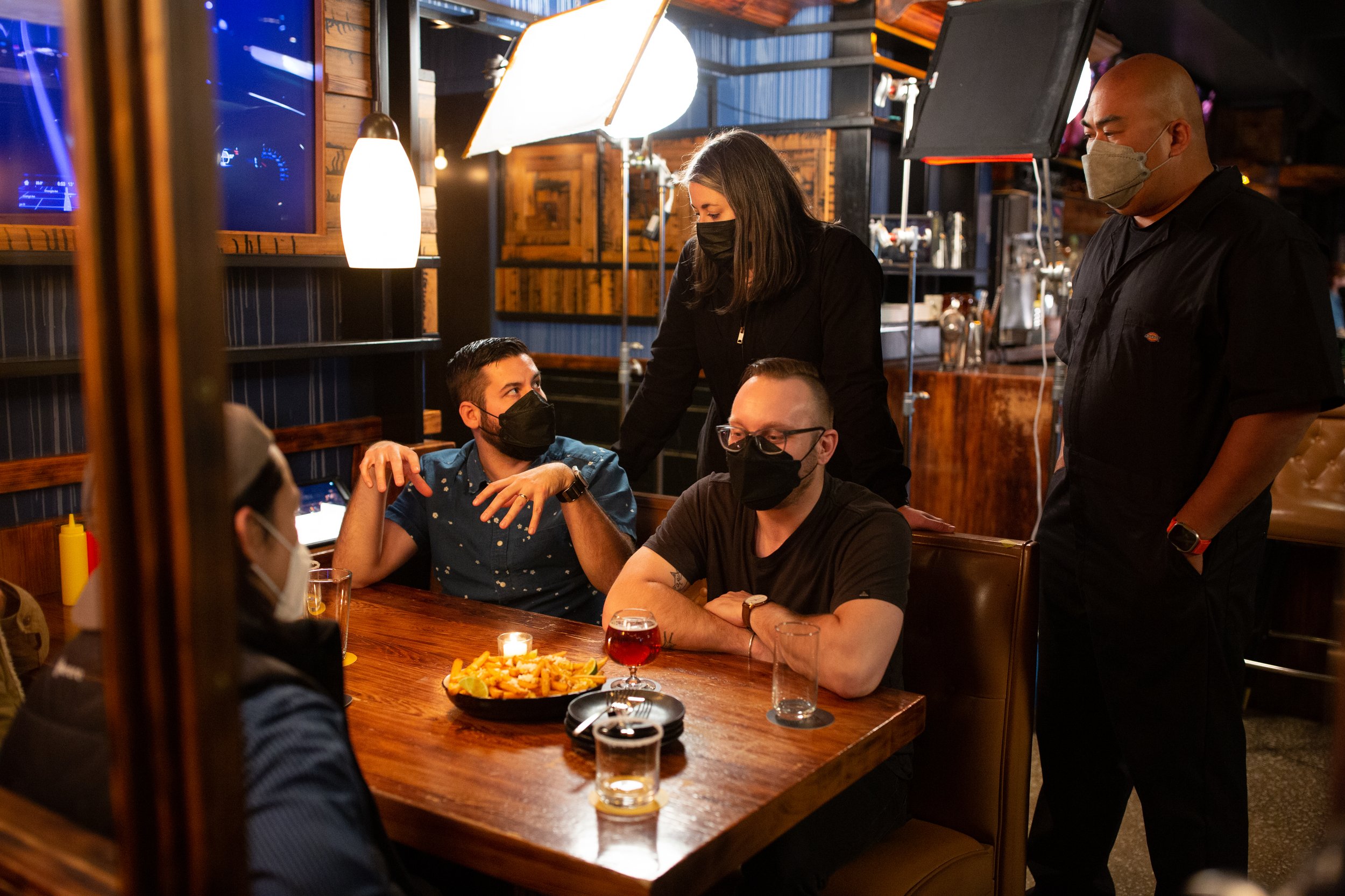 Group of people wearing masks seated at a restaurant table having a conversation, with food and drinks on the table, and lighting equipment in the background.