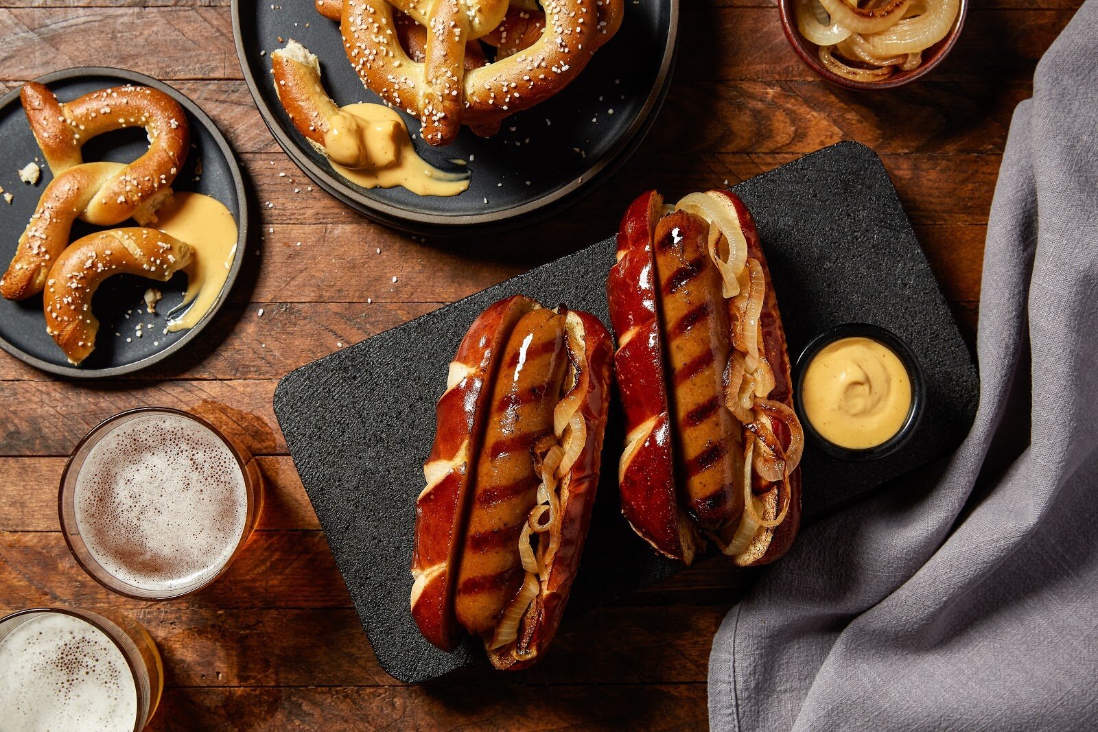 Grilled sausages in pretzel buns with caramelized onions and mustard, accompanied by soft pretzels with cheese sauce, and pints of beer on a wooden table.