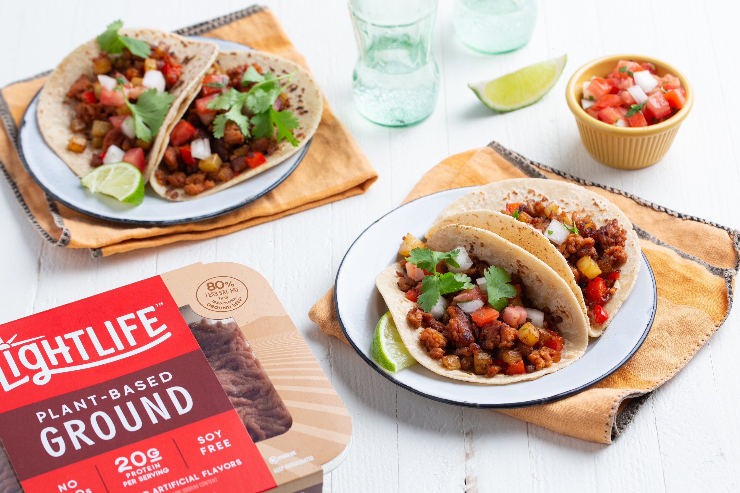 Tacos with plant-based filling, topped with diced tomatoes, onions, and cilantro, served on white plates with lime wedges. A package of Lightlife plant-based ground meat is in the foreground. A small bowl of salsa and glass bottles are in the backgro