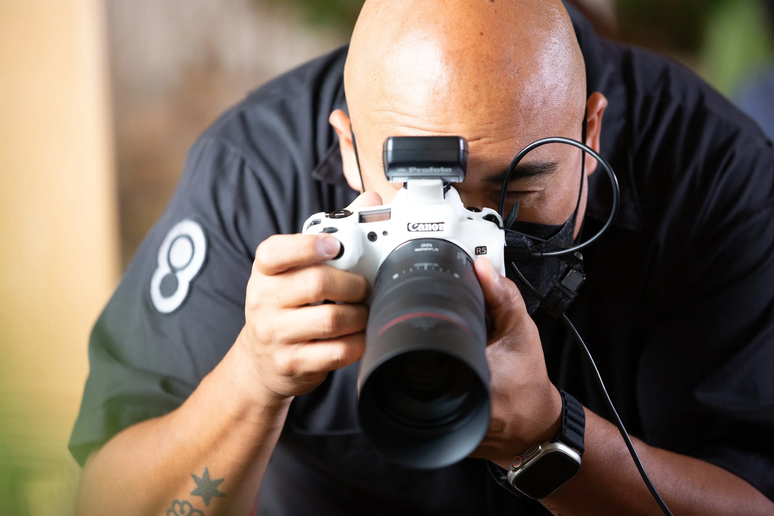 Photographer holding a Canon R5 camera with a large lens, wearing a mask, and focusing on taking a picture.